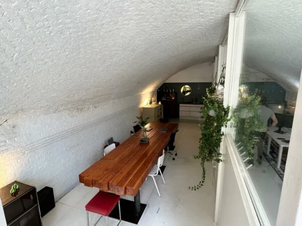 Vergaderruimte onder een witgewelfd plafond met een lange houten tafel, stoelen en uitzicht op een aangrenzend kantoor via een raam.